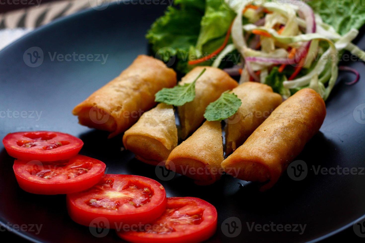 Fried spring rolls with vegetables and tomatoes placed in a black plate on a black wooden table and dipping sauce. photo