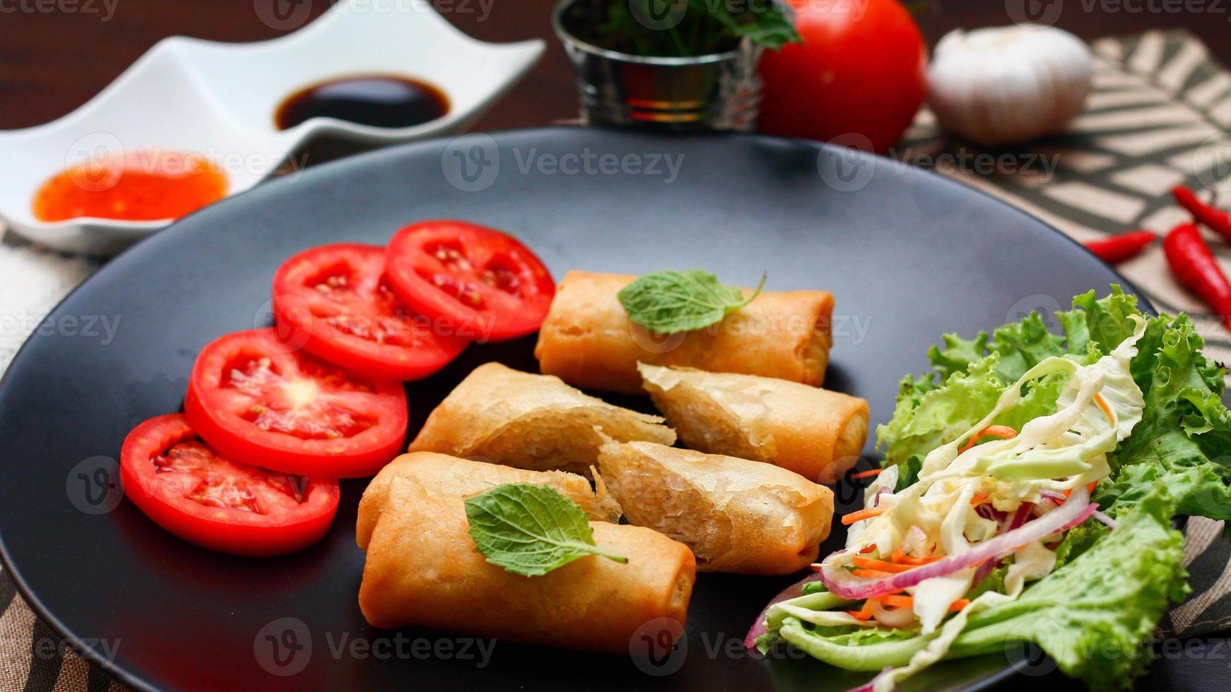 Fried spring rolls with vegetables and tomatoes placed in a black plate on a black wooden table and dipping sauce. photo