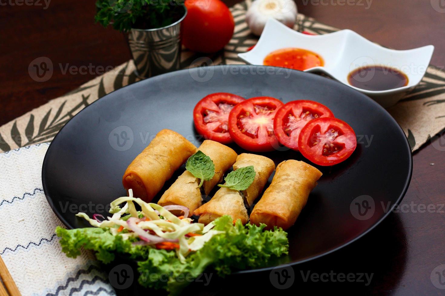 Fried spring rolls with vegetables and tomatoes placed in a black plate on a black wooden table and dipping sauce. photo