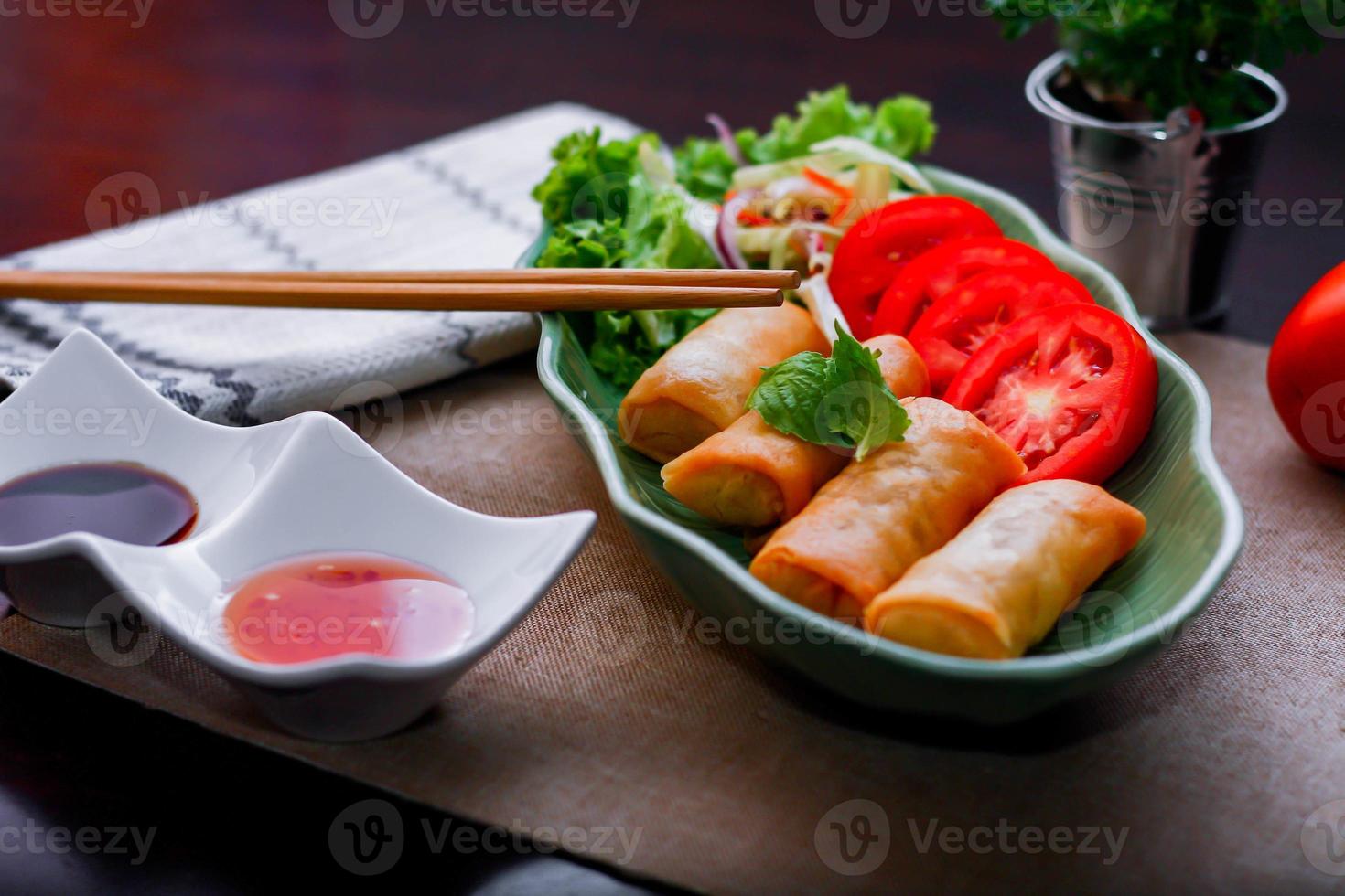 Fried spring rolls, vegetables and tomatoes placed in a green leaf shape plate on a black wooden table and dipping sauce. photo