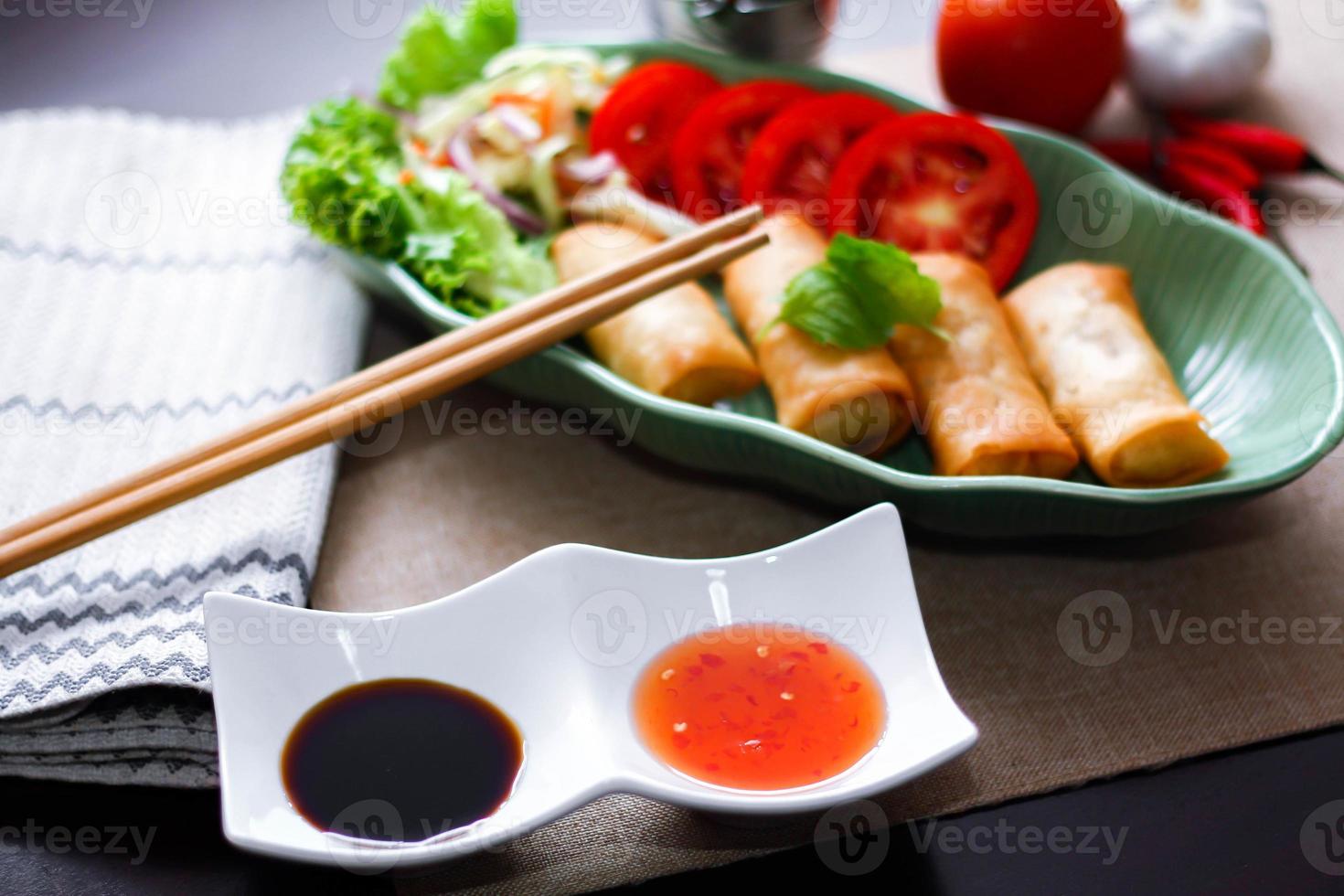 Fried spring rolls, vegetables and tomatoes placed in a green leaf shape plate on a black wooden table and dipping sauce. photo