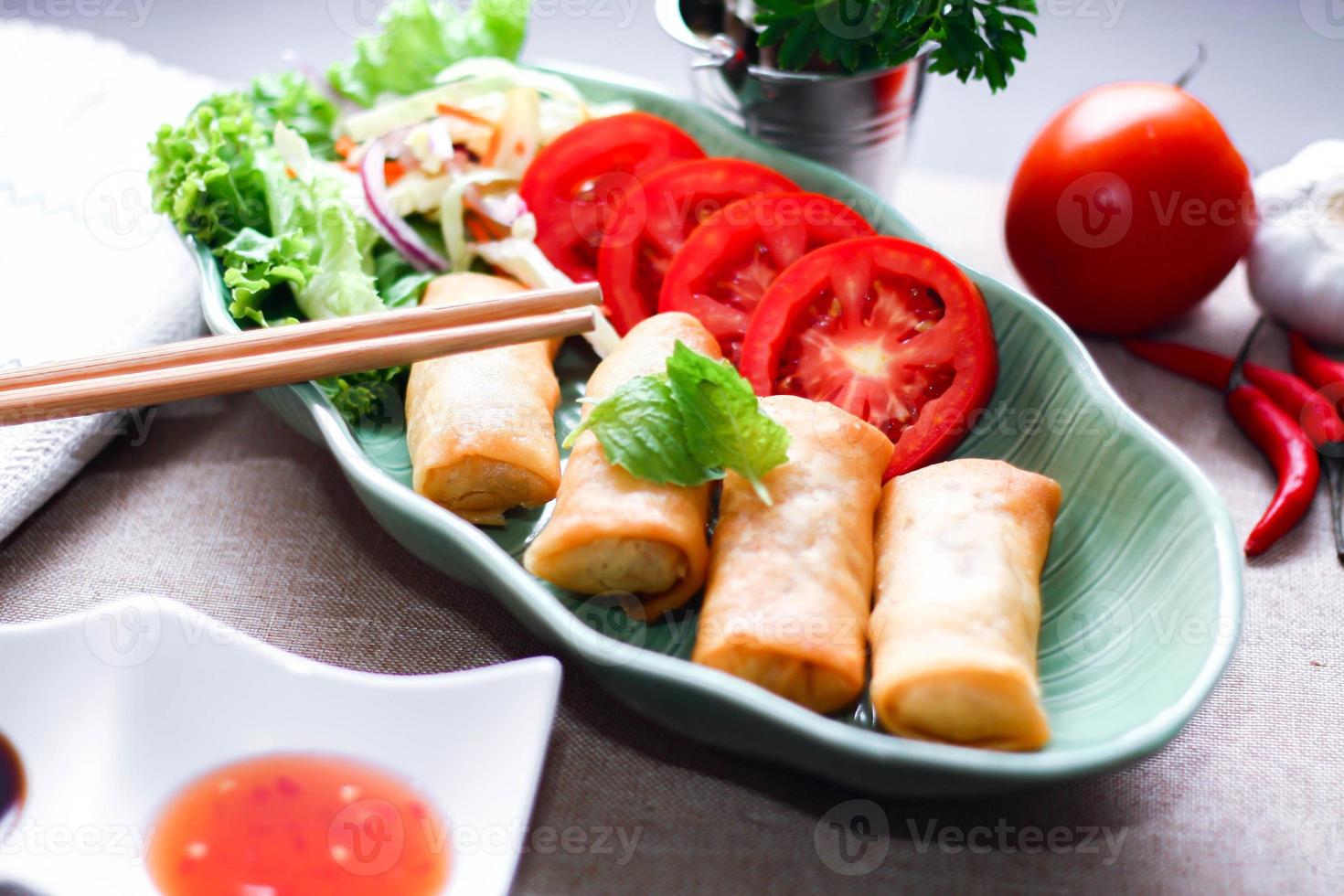 Fried spring rolls, vegetables and tomatoes placed in a green leaf shape plate on a black wooden table and dipping sauce. photo