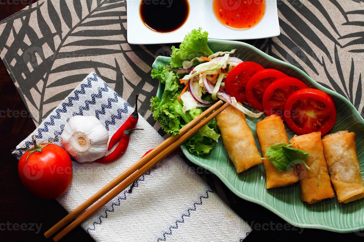 Fried spring rolls, vegetables and tomatoes placed in a green leaf shape plate on a black wooden table and dipping sauce. photo