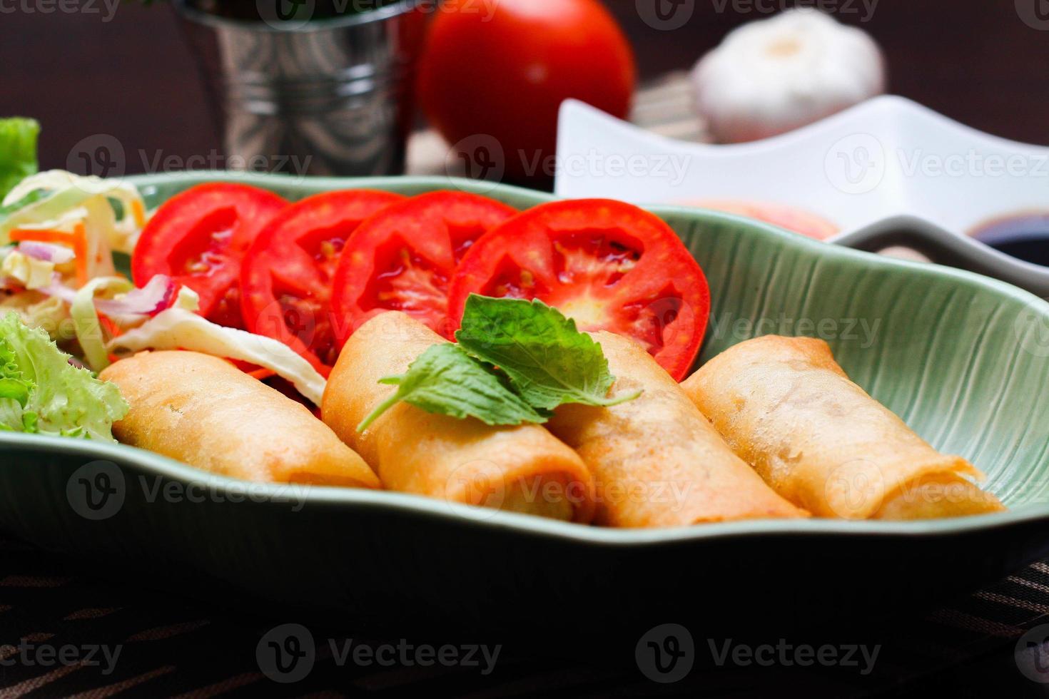 Fried spring rolls, vegetables and tomatoes placed in a green leaf shape plate on a black wooden table and dipping sauce. photo