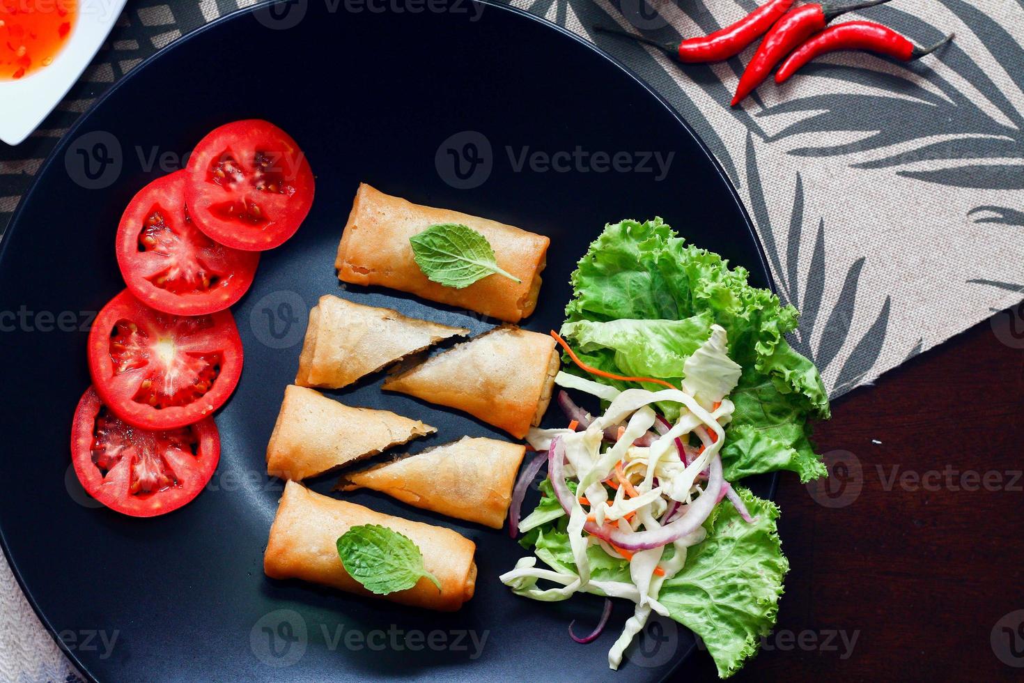 Fried spring rolls with vegetables and tomatoes placed in a black plate on a black wooden table and dipping sauce. photo