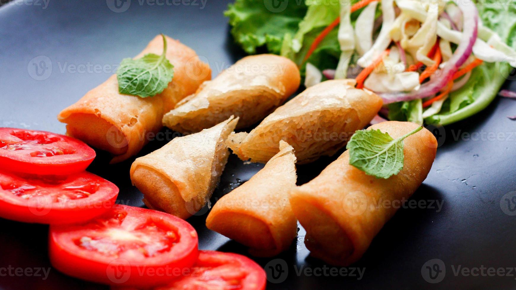 Fried spring rolls with vegetables and tomatoes placed in a black plate on a black wooden table and dipping sauce. photo