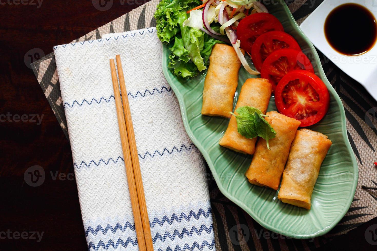 Fried spring rolls, vegetables and tomatoes placed in a green leaf shape plate on a black wooden table and dipping sauce. photo