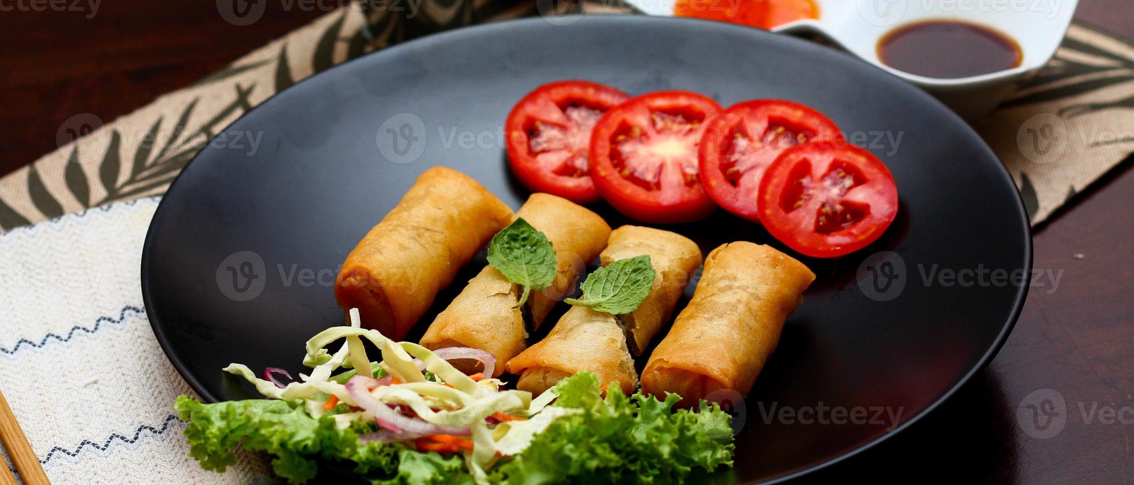 Fried spring rolls with vegetables and tomatoes placed in a black plate on a black wooden table and dipping sauce. photo
