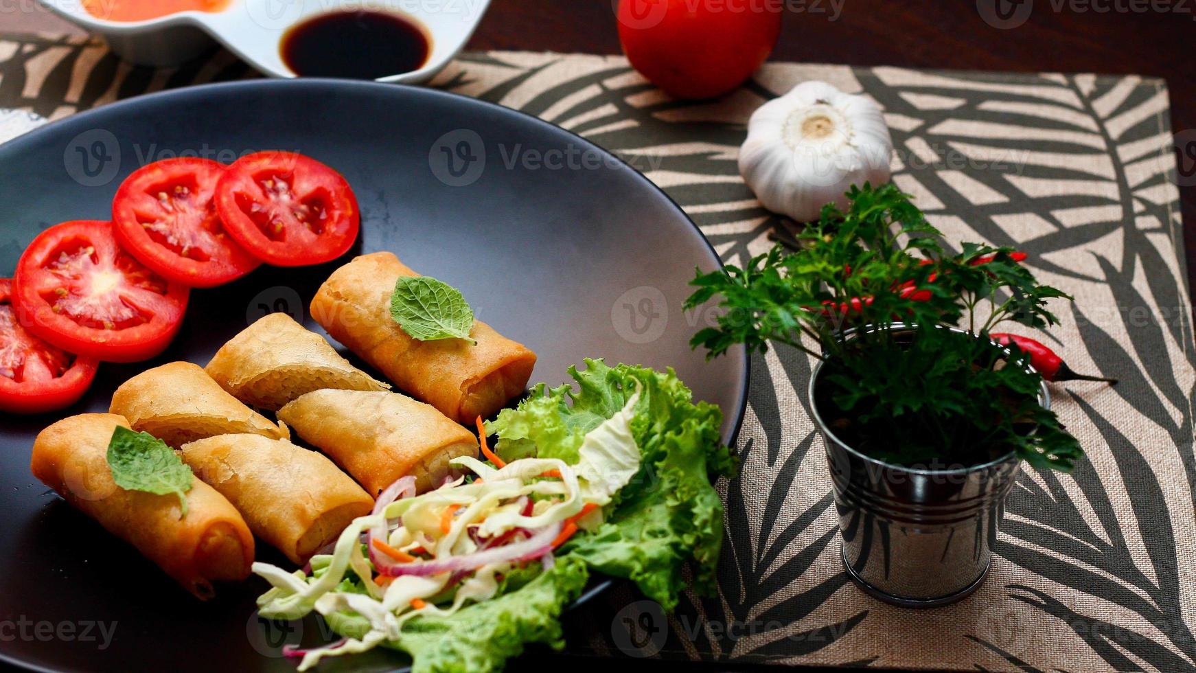 Fried spring rolls with vegetables and tomatoes placed in a black plate on a black wooden table and dipping sauce. photo