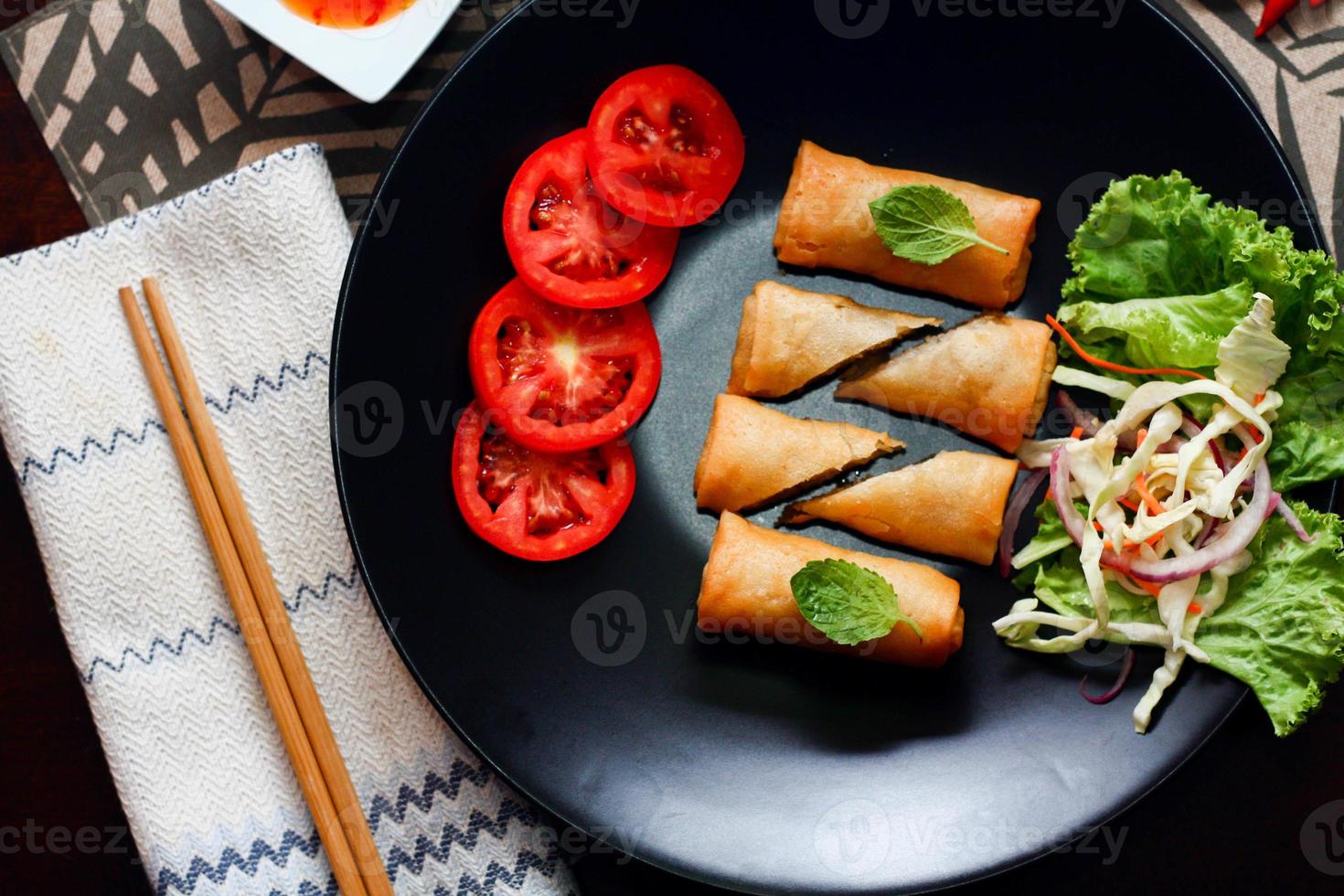 Fried spring rolls with vegetables and tomatoes placed in a black plate on a black wooden table and dipping sauce. photo