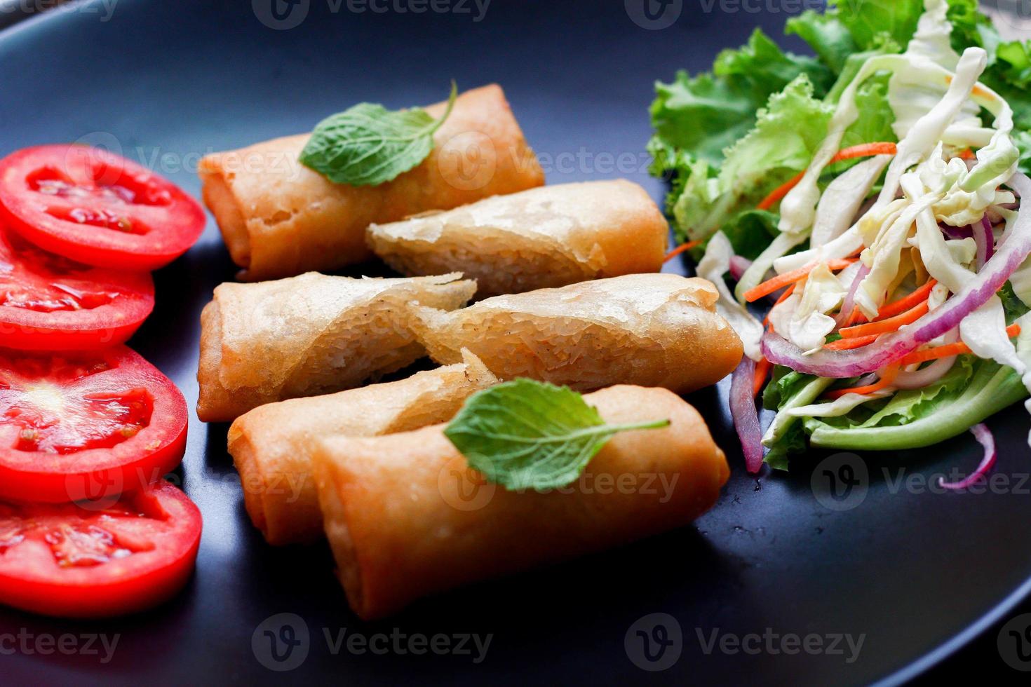 Fried spring rolls with vegetables and tomatoes placed in a black plate on a black wooden table and dipping sauce. photo