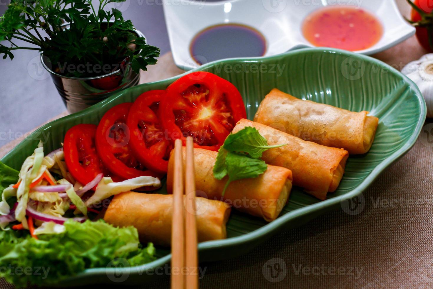 Fried spring rolls, vegetables and tomatoes placed in a green leaf shape plate on a black wooden table and dipping sauce. photo