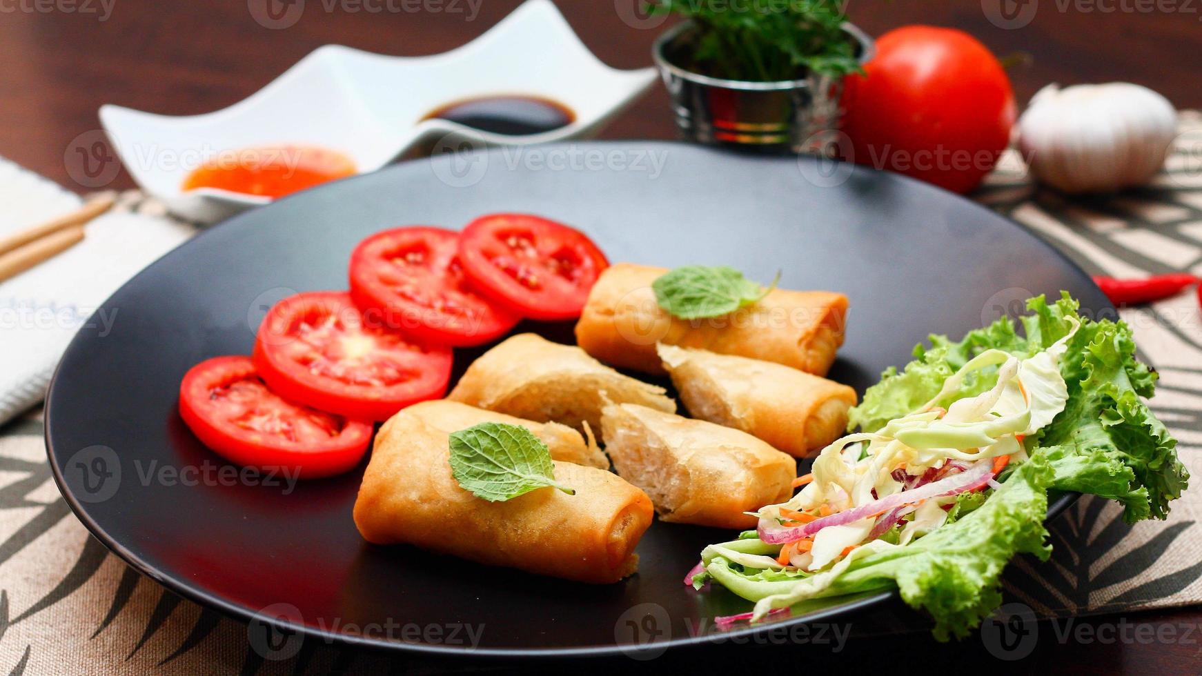 Fried spring rolls with vegetables and tomatoes placed in a black plate on a black wooden table and dipping sauce. photo