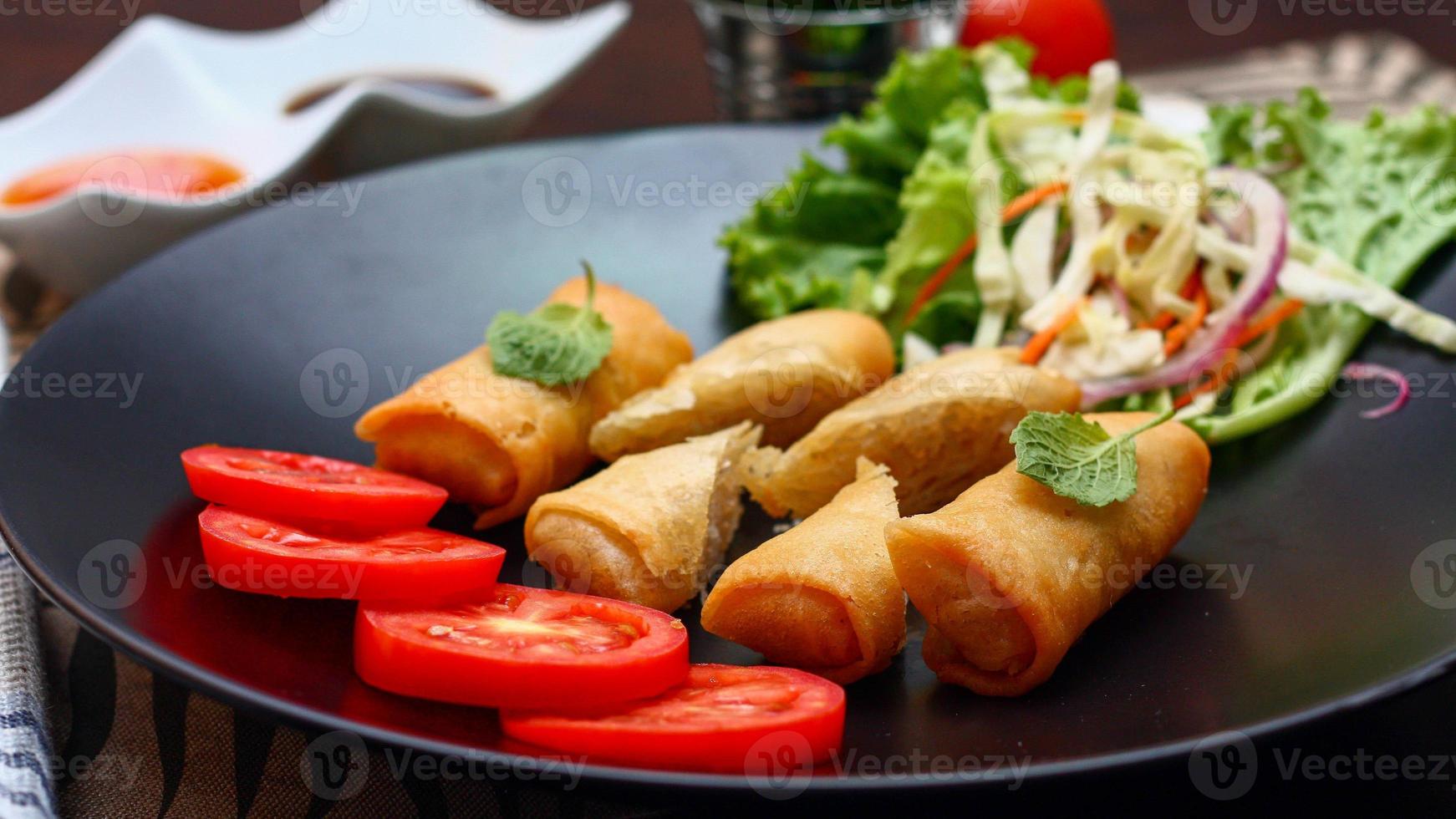 frito primavera rollos con vegetales y Tomates metido en un negro plato en un negro de madera mesa y inmersión salsa. foto