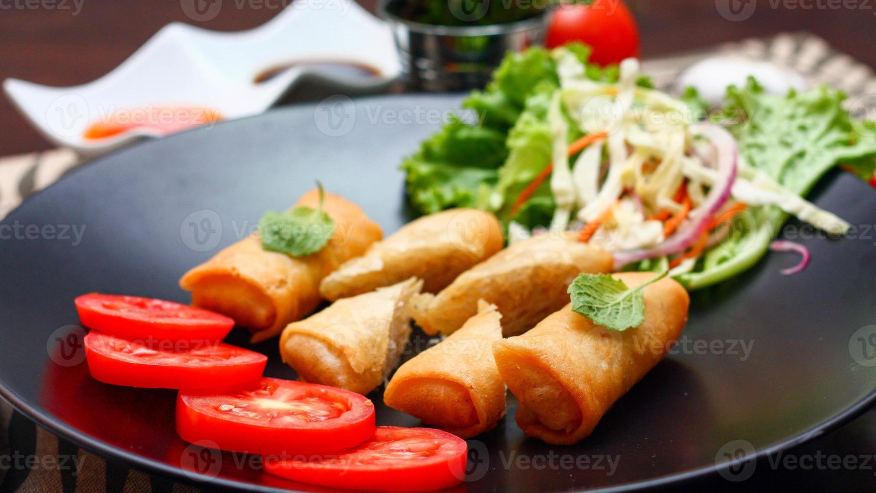 Fried spring rolls with vegetables and tomatoes placed in a black plate on a black wooden table and dipping sauce. photo