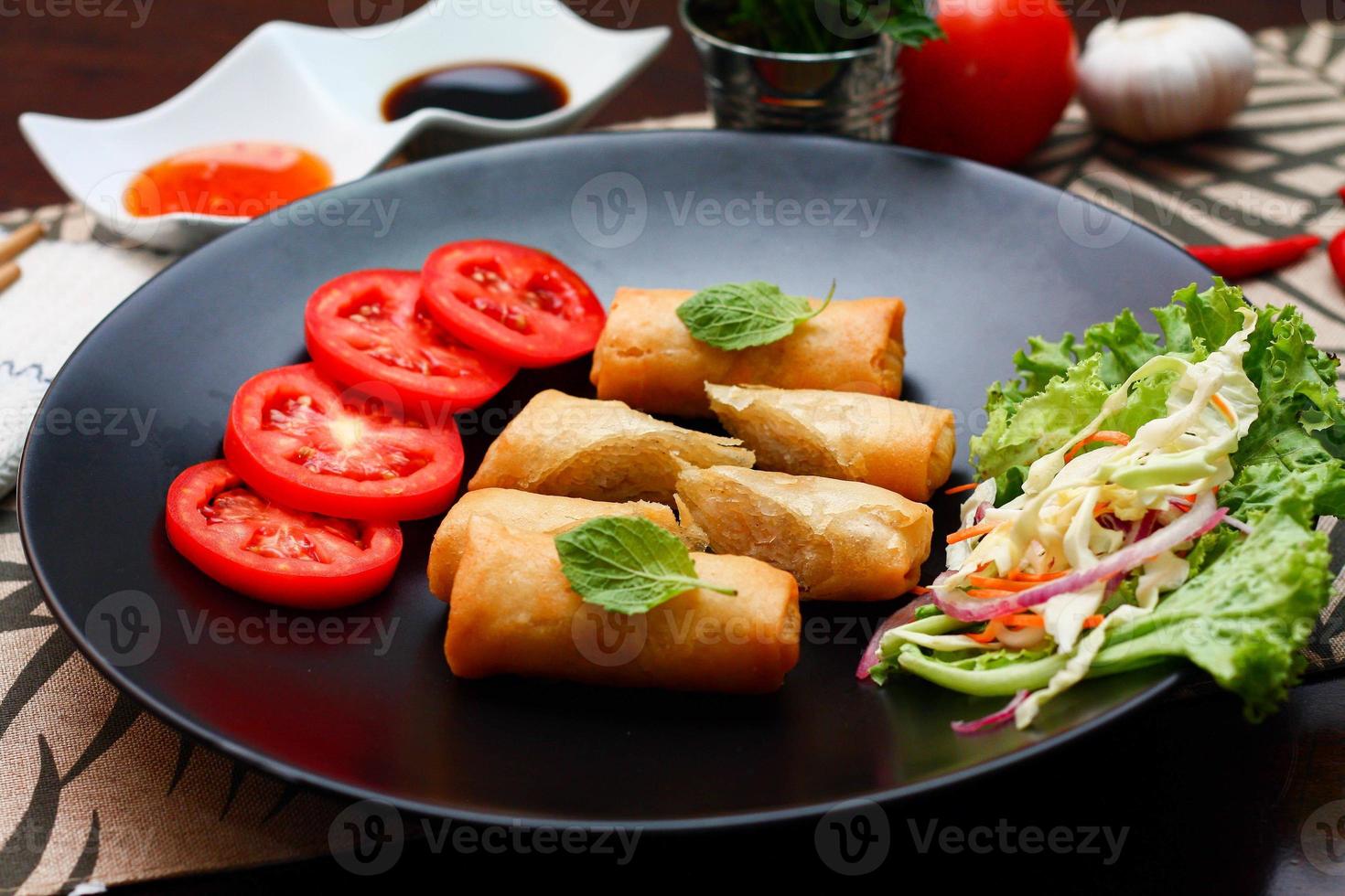 Fried spring rolls with vegetables and tomatoes placed in a black plate on a black wooden table and dipping sauce. photo