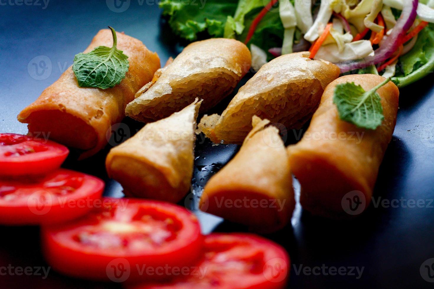 Fried spring rolls with vegetables and tomatoes placed in a black plate on a black wooden table and dipping sauce. photo