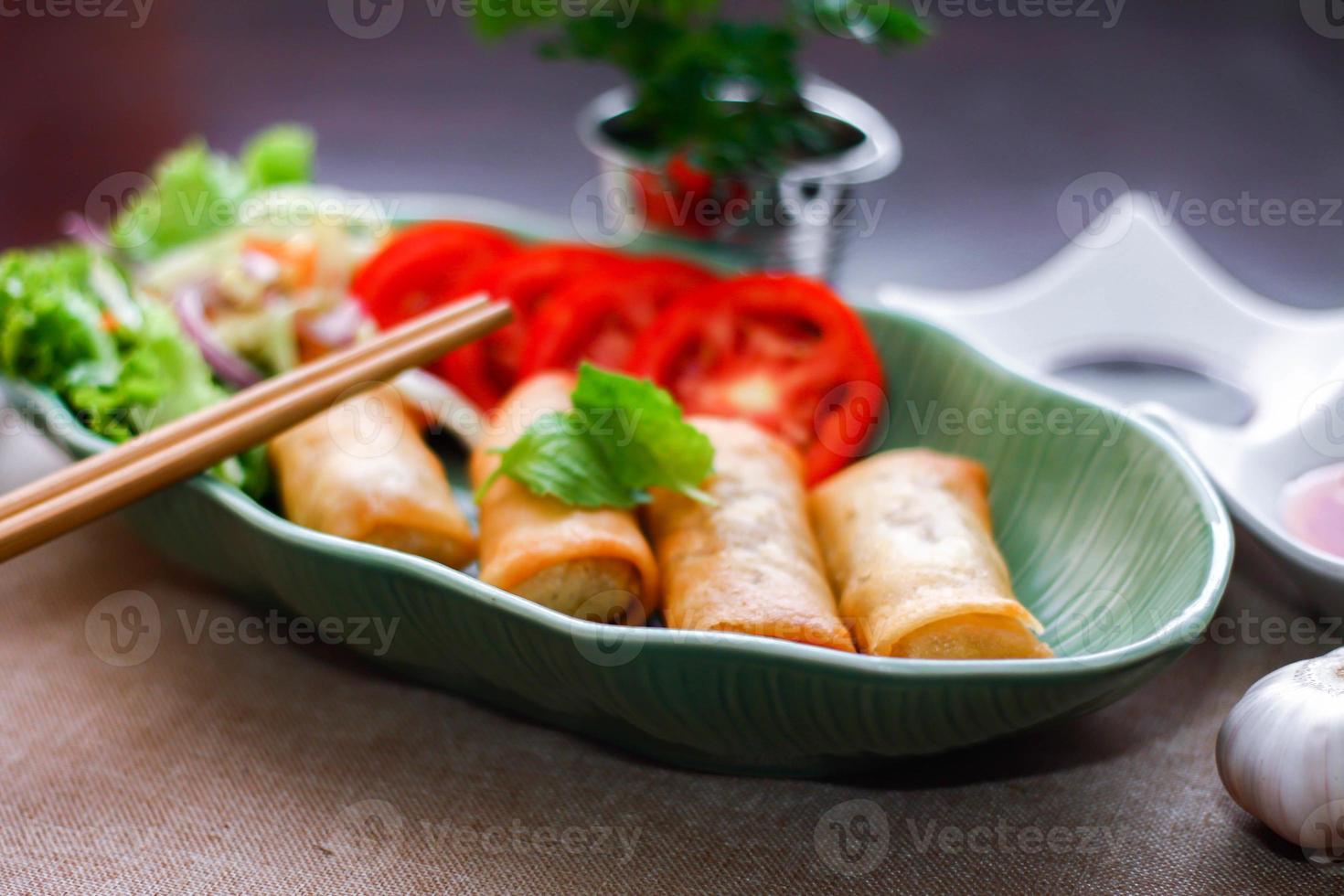 Fried spring rolls, vegetables and tomatoes placed in a green leaf shape plate on a black wooden table and dipping sauce. photo