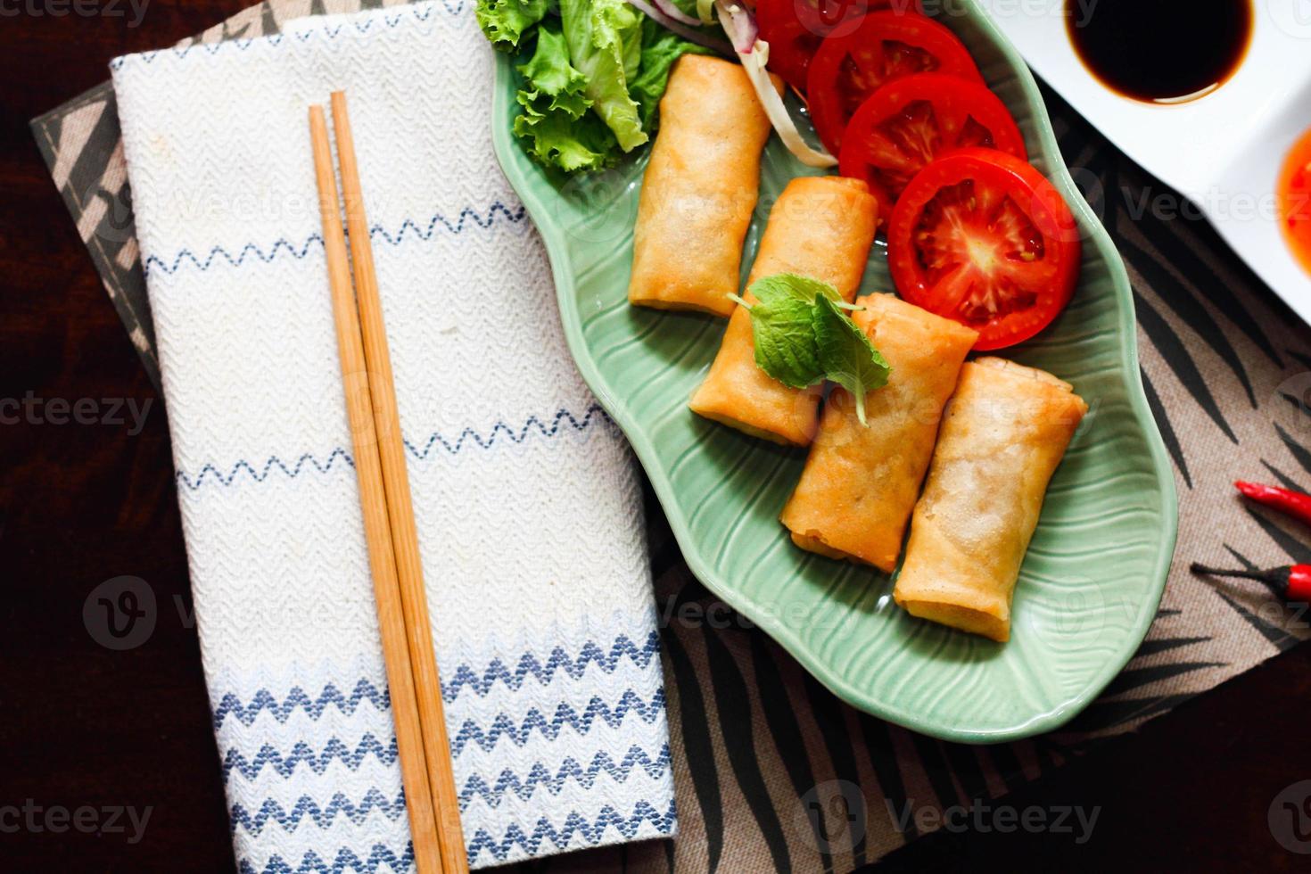 Fried spring rolls, vegetables and tomatoes placed in a green leaf shape plate on a black wooden table and dipping sauce. photo