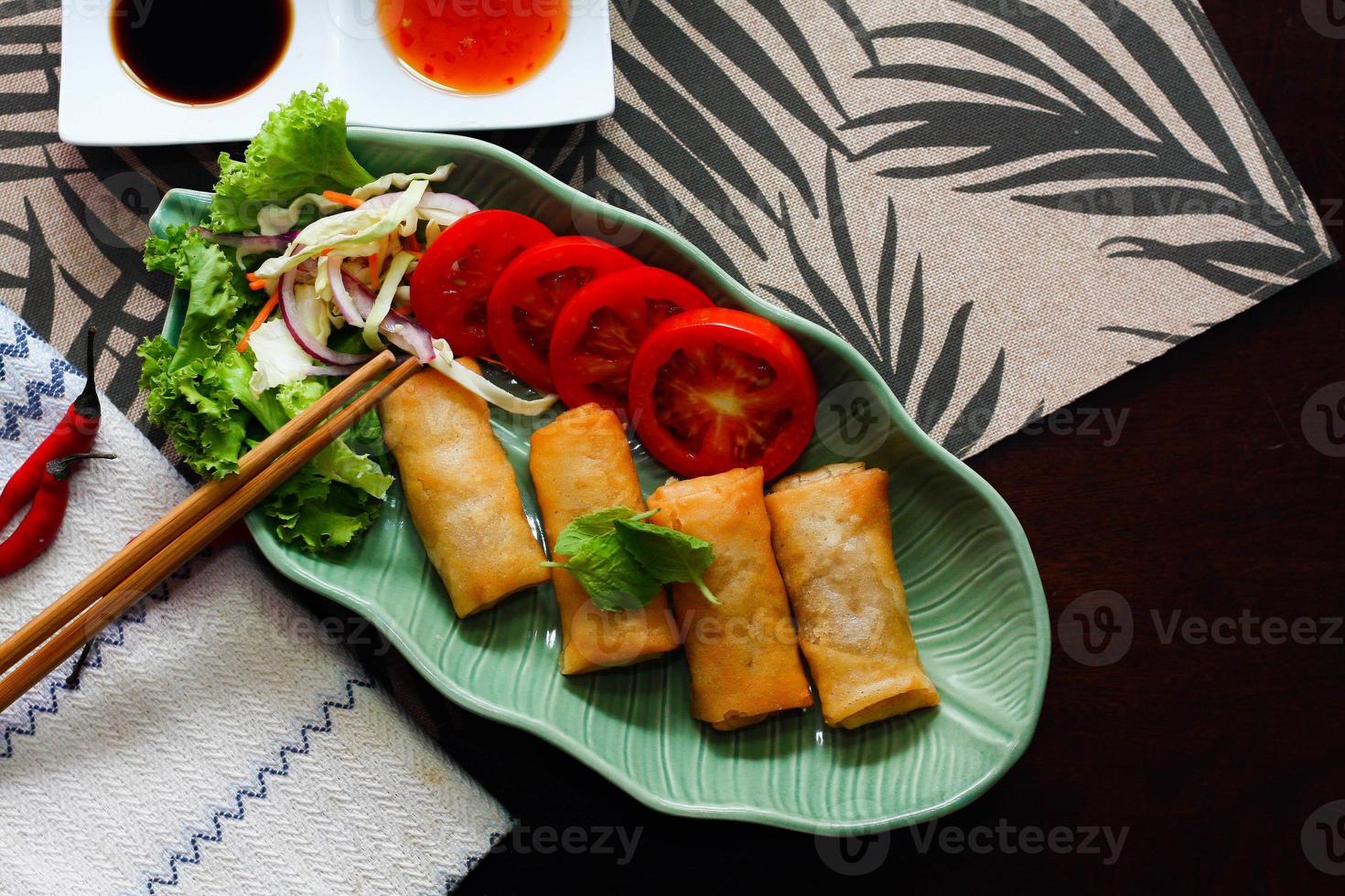 Fried spring rolls, vegetables and tomatoes placed in a green leaf shape plate on a black wooden table and dipping sauce. photo