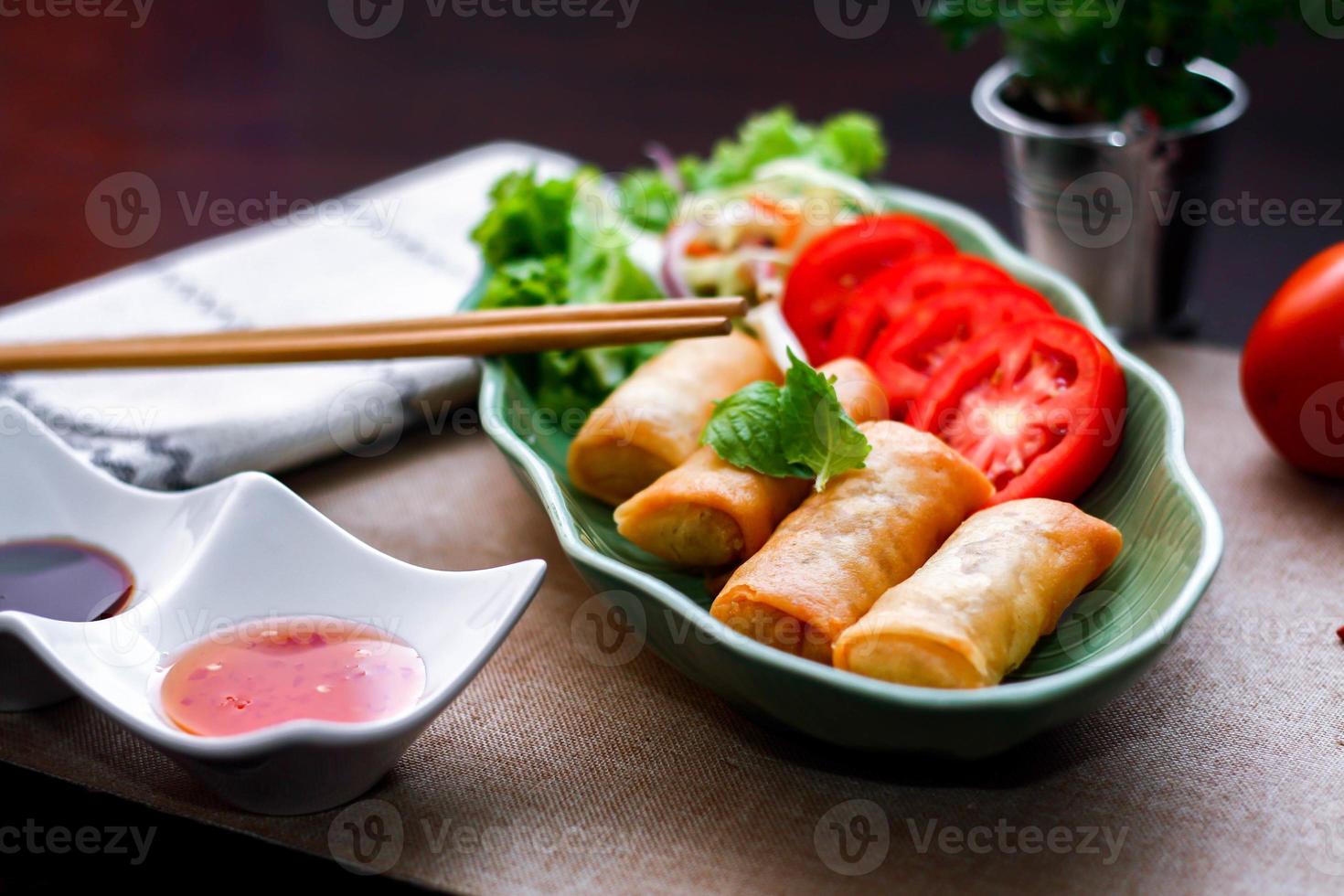 Fried spring rolls, vegetables and tomatoes placed in a green leaf shape plate on a black wooden table and dipping sauce. photo