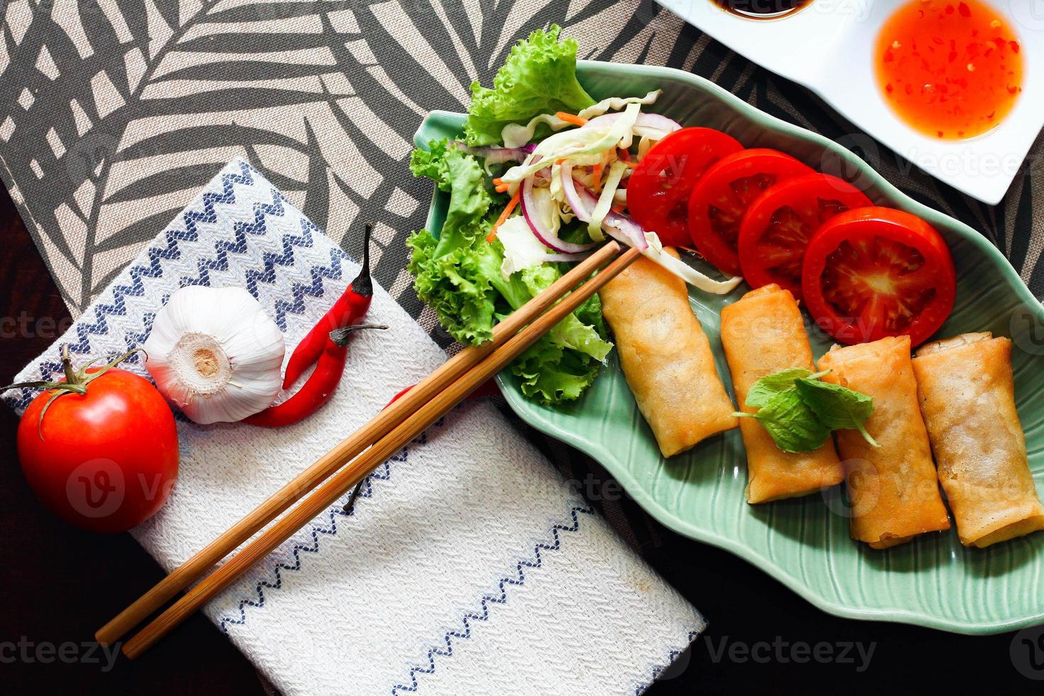 Fried spring rolls, vegetables and tomatoes placed in a green leaf shape plate on a black wooden table and dipping sauce. photo