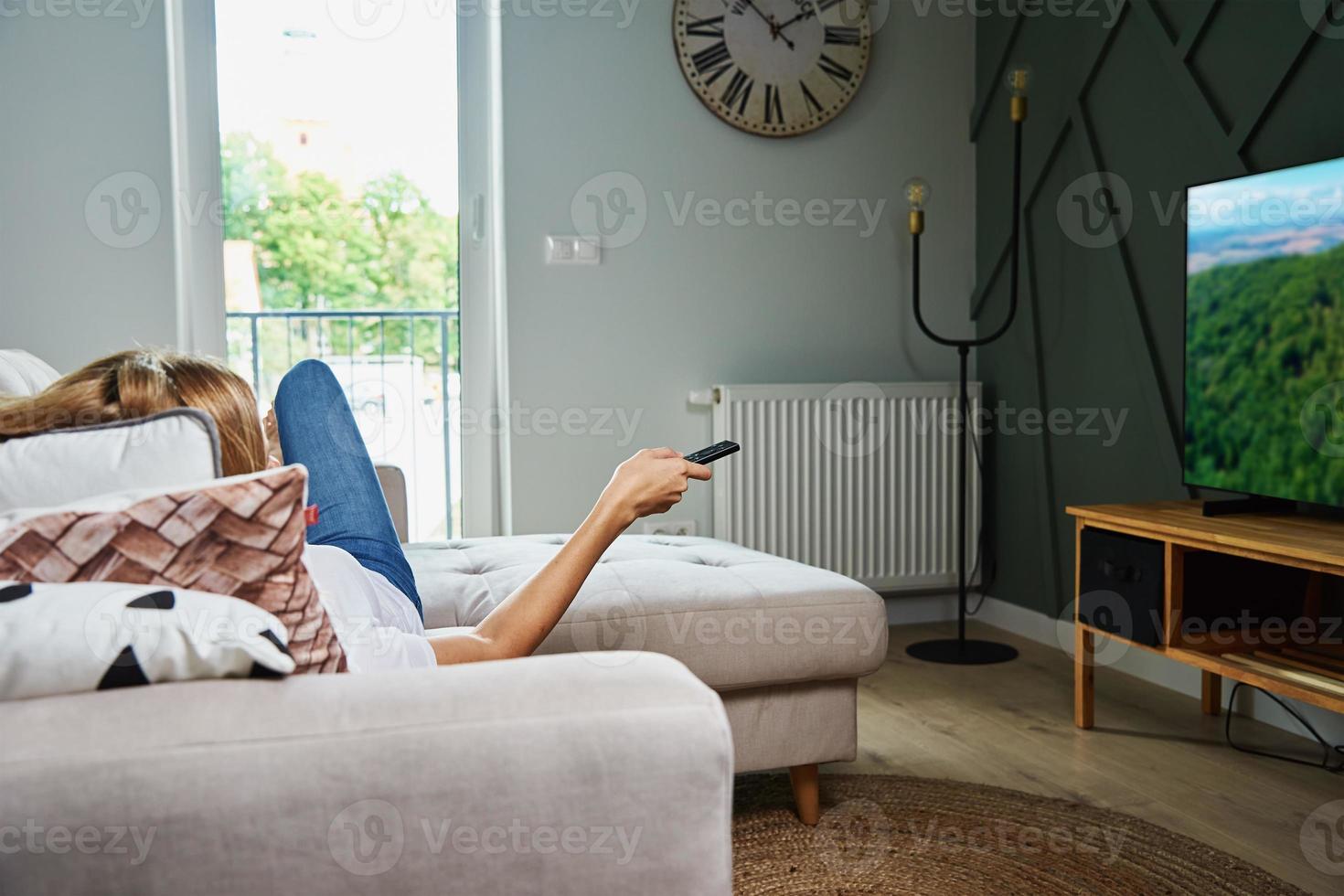Woman watch TV set while luying on the sofa and switch channels with remote control photo