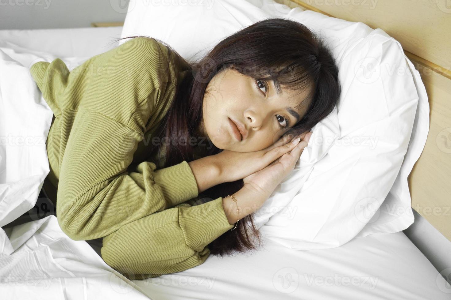 Thoughtful Asian woman looks pensively above dressed in sweatshirt poses thinks about future while lying on the bed photo