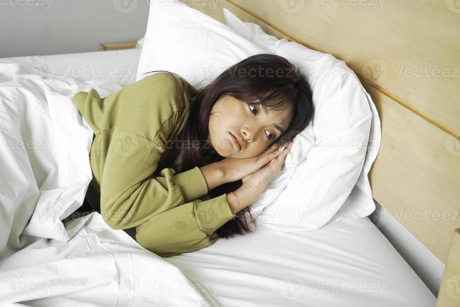 Thoughtful Asian woman looks pensively above dressed in sweatshirt poses thinks about future while lying on the bed photo