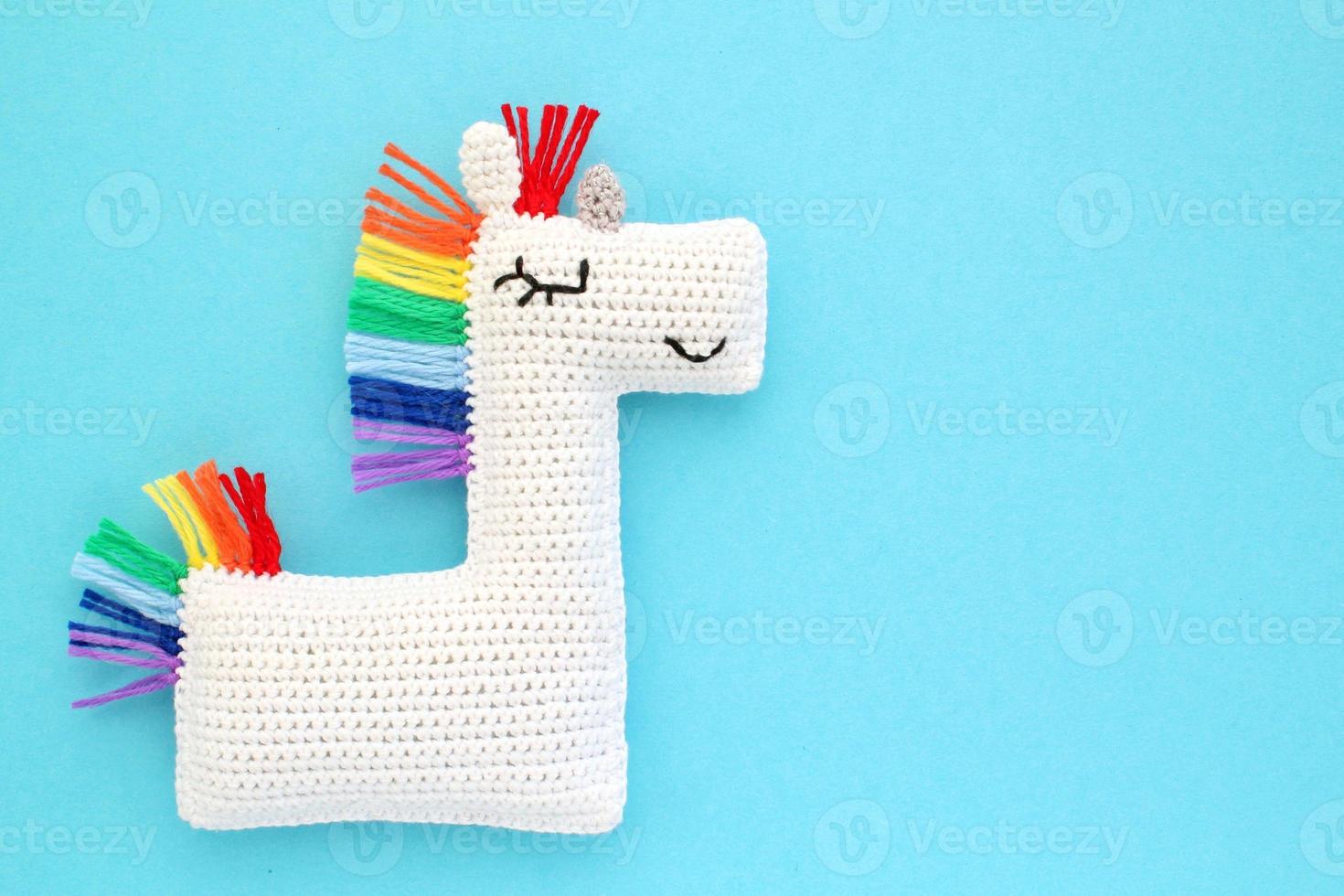 Crochet amigurumi handmade stuffed soft white unicorn toy with rainbow mane on blue background. Handwork, hobby. Craft diy newborn pregnancy concept. Knitted doll for little baby. Close up flat lay photo