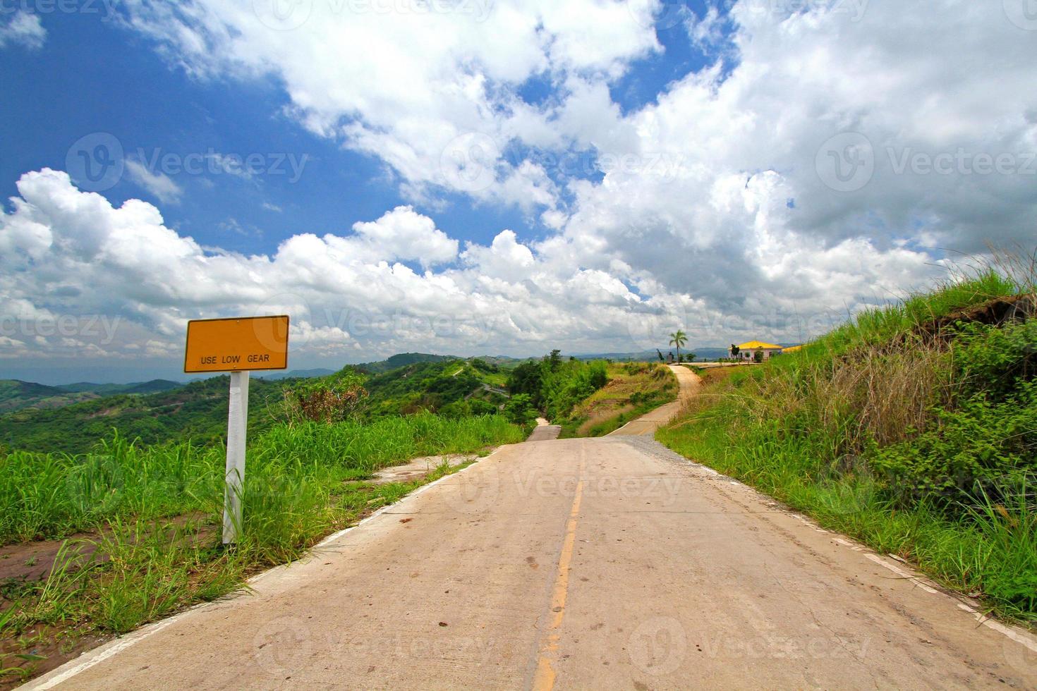 The sign tell tourist the road is slope and use low gear for driving. Road among beautiful in nature with grass, blue sky and clouds background. Travel to destination photo