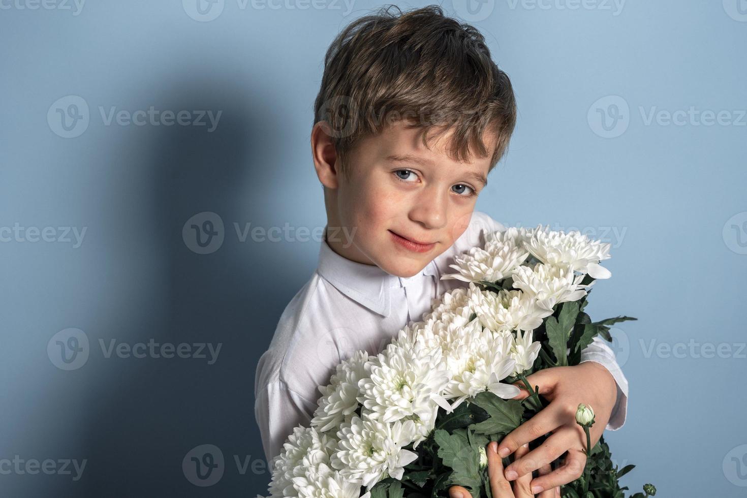 A cute Caucasian boy in a white shirt holds a bouquet of white flowers in his hands and smiles. photo