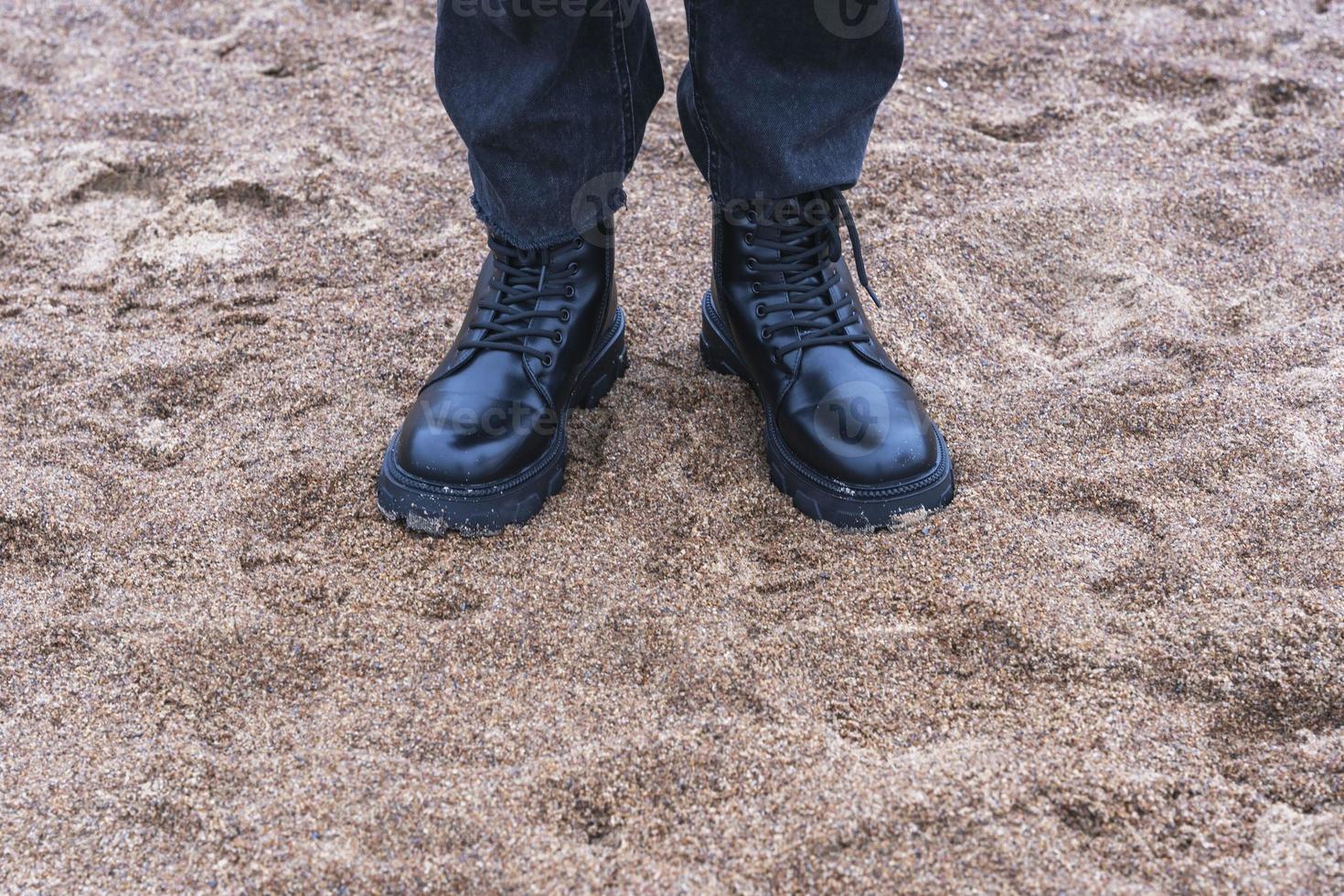 Feet in black leather rough boots and black fashionable denim pants standing on the sand on the seashore photo