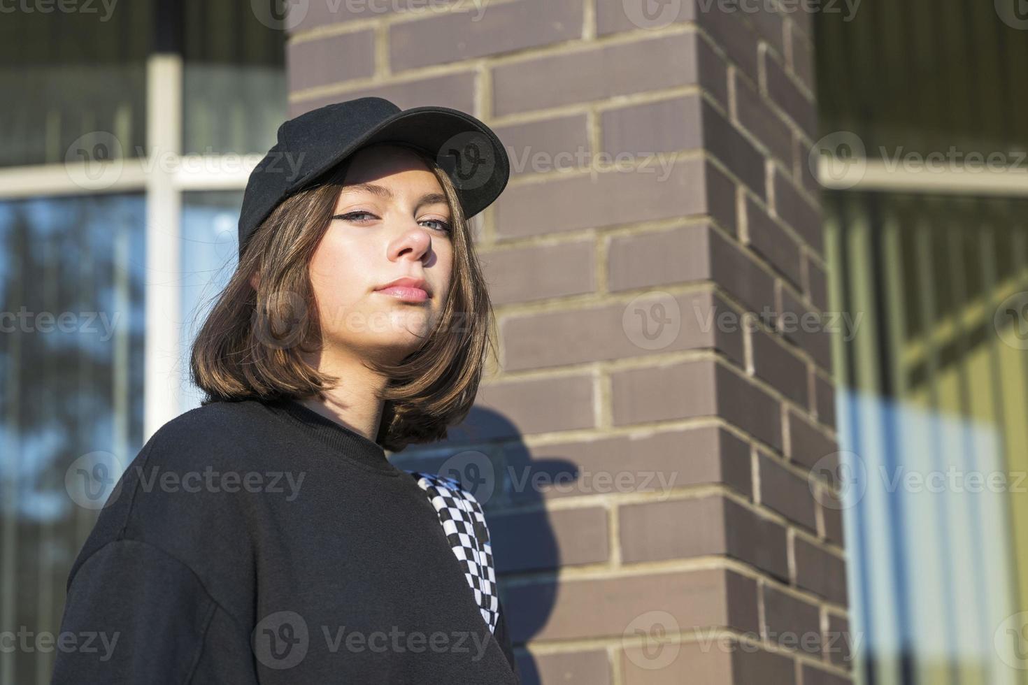 linda caucásico niña con un mochila, vestido en un de moda negro béisbol gorra y un negro capucha foto