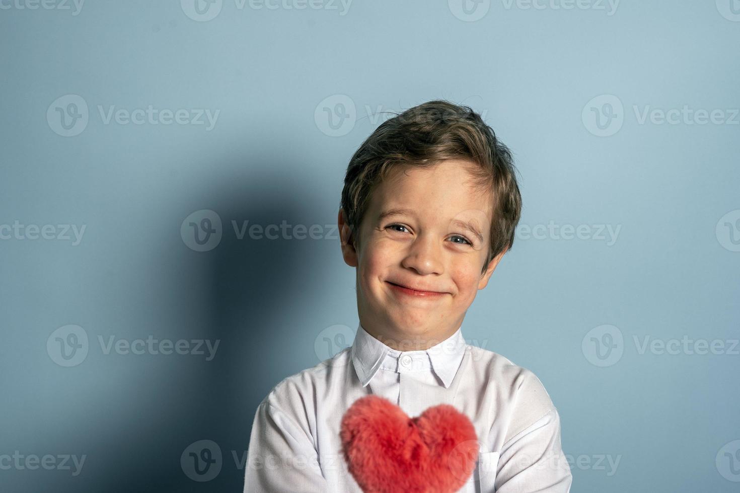 A cute Caucasian boy in a white shirt holds a fluffy red heart in his hands photo