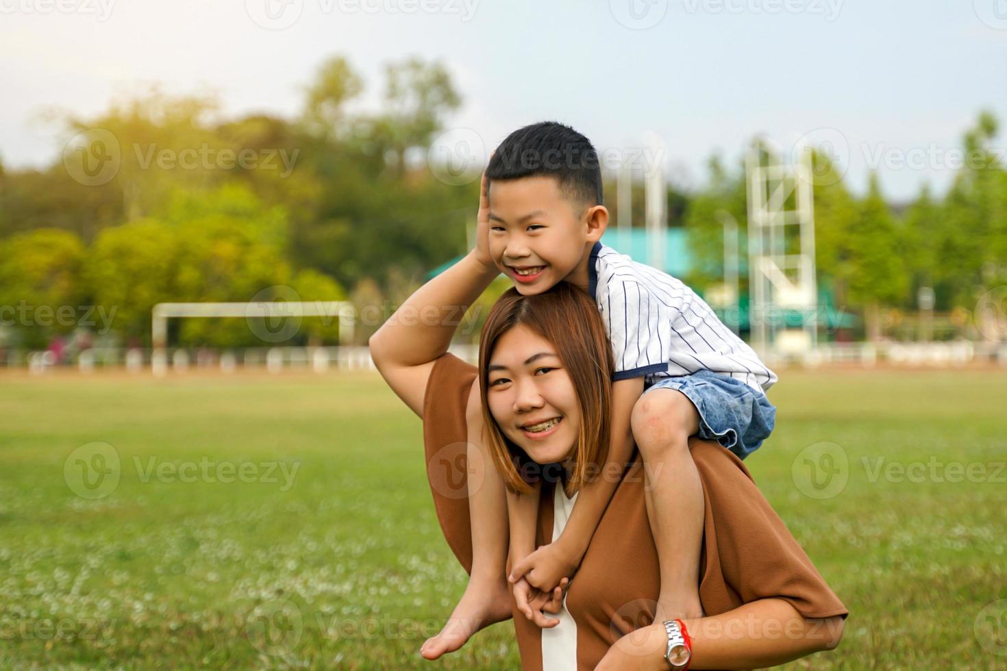 Son riding on mother's back, laughing and playing happily on the outdoor lawn. Soft and selective focus. photo