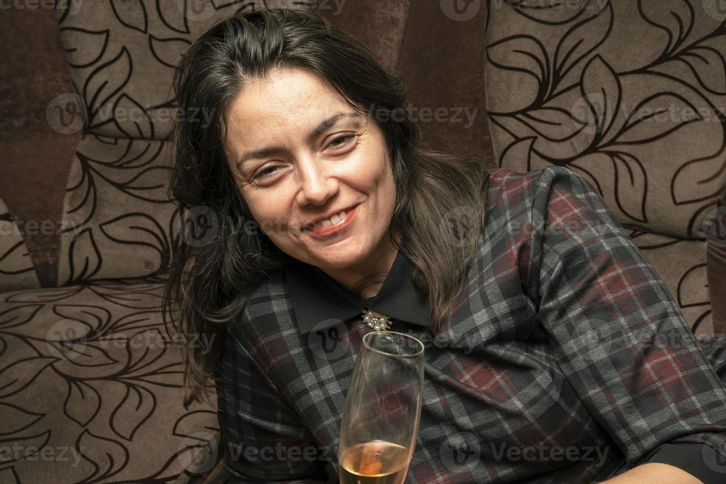 A young beautiful woman in a dress drinks wine from a glass lying on a home sofa and smiles photo