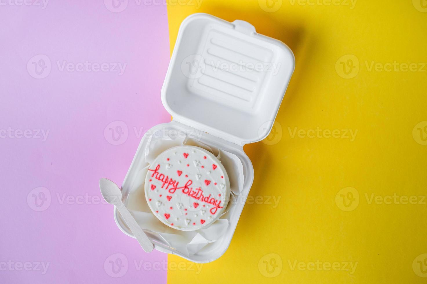 A small bento birthday cake in a box. Wooden spoon with white ribbon. Yellow and pink background photo