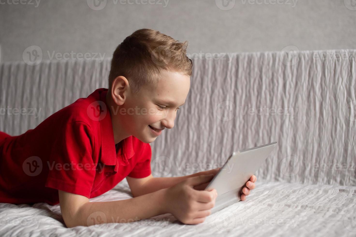 A cute boy in a red T-shirt is lying on the couch with a tablet and laughing photo