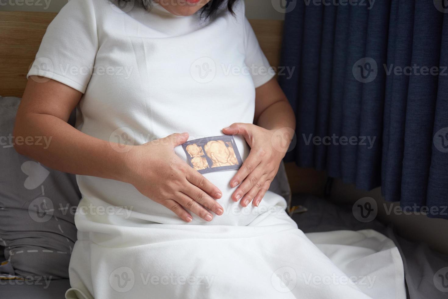 Image of Asian pregnant smiling woman sitting and posing while showing ultrasound scans, look into the camera photo