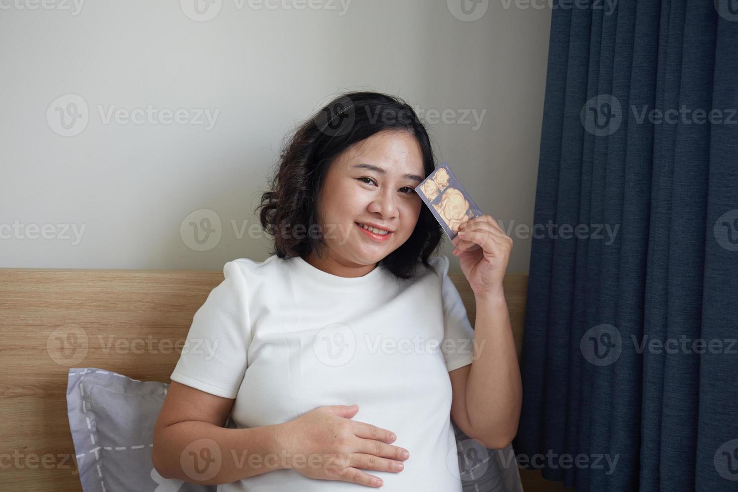 Image of Asian pregnant smiling woman sitting and posing while showing ultrasound scans, look into the camera photo