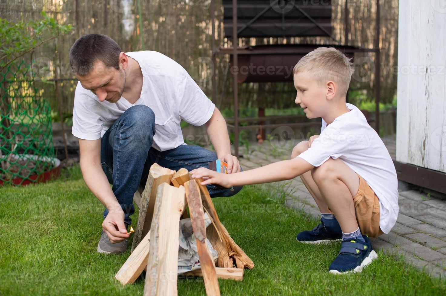 A cute boy in a white T-shirt is building a place for a bonfire next to his dad photo