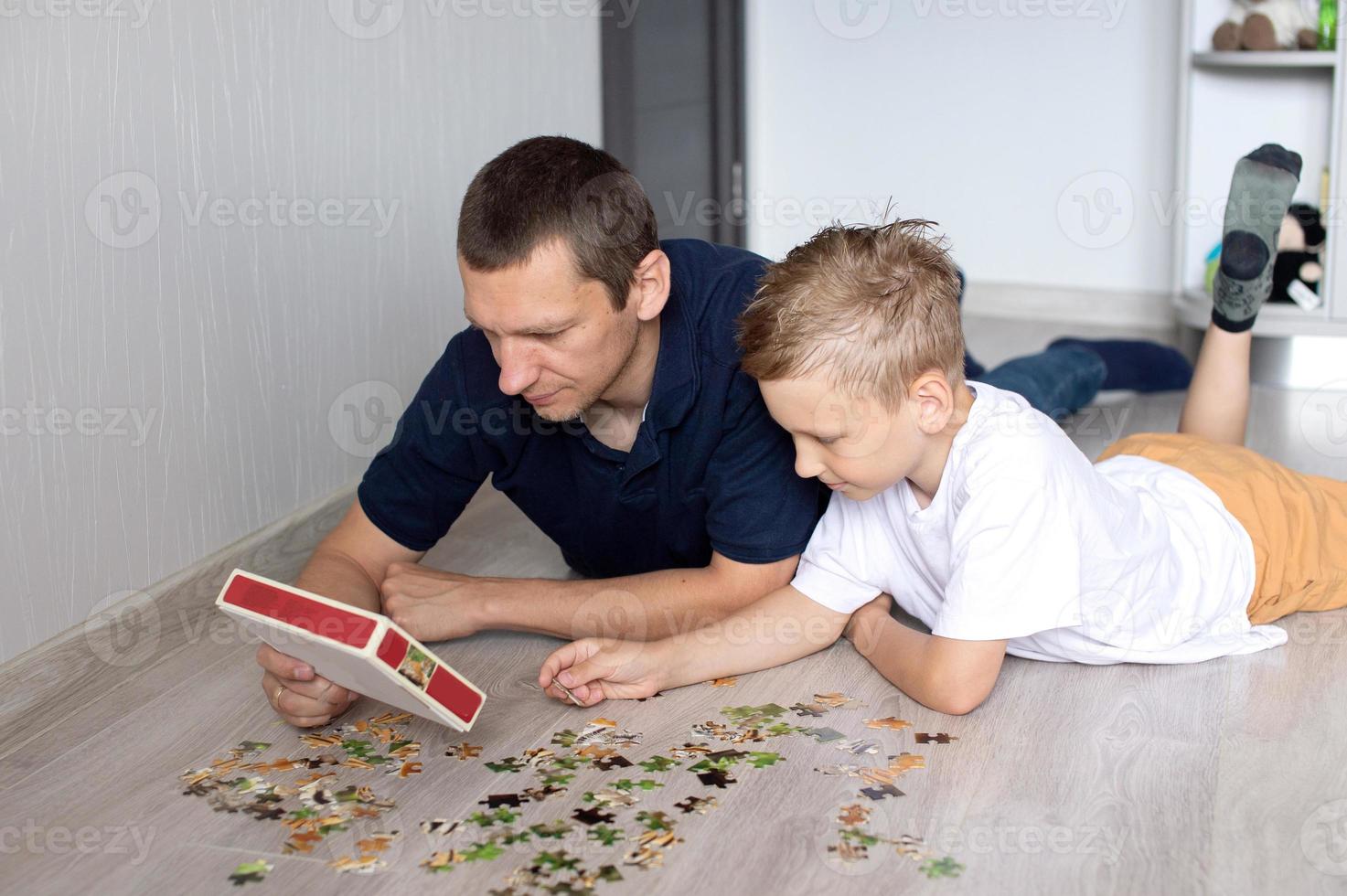 A cute boy with a cheerful dad is collecting a puzzle lying on the floor in a room photo