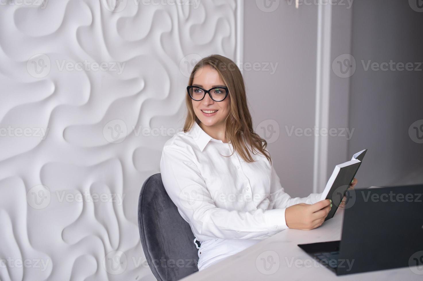 A young teacher with glasses holds a diary in her hands and sits at a table with a laptop photo