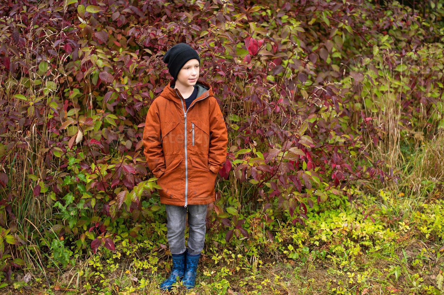 A cute boy in rubber boots stands in the autumn forest photo