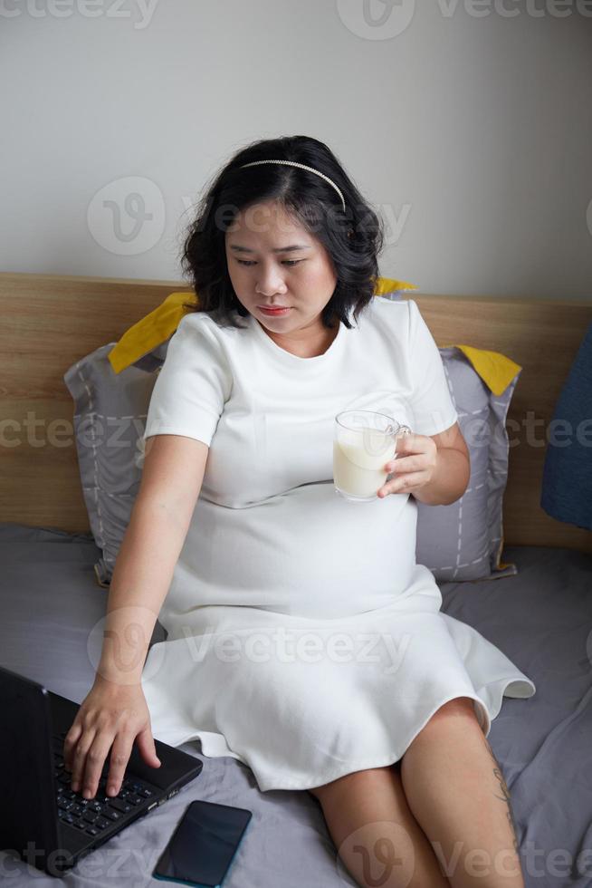 Happy Young Asian Pregnant Woman drinking milk on bed at home while holding her belly and thinking photo