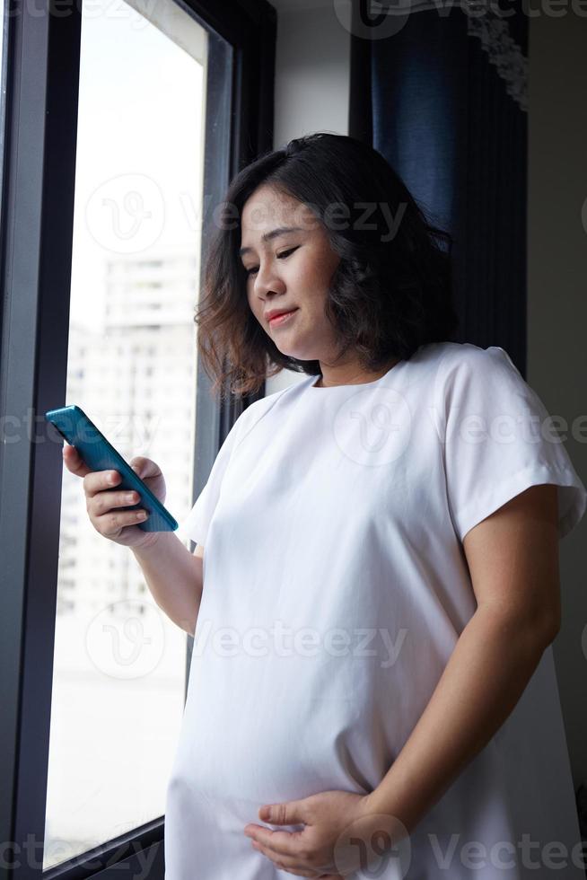Young Asian Pregnant Woman standing near the window and using smart phone for checking information photo
