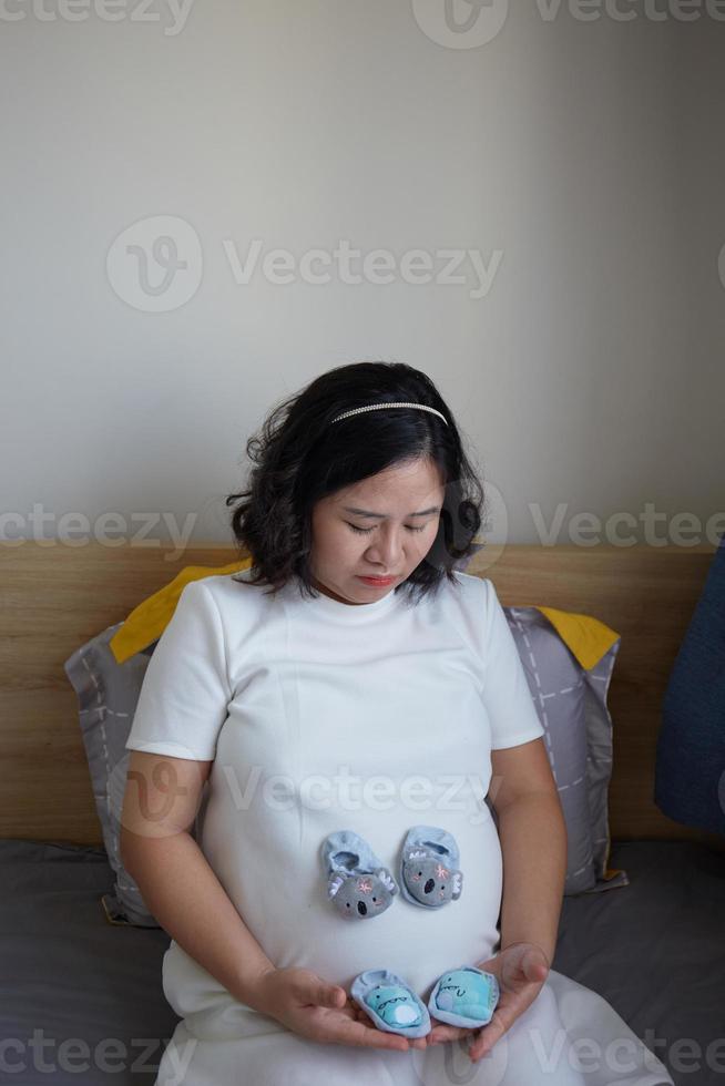 Photo of Happy Asian pregnant woman sitting in bed and touching her belly at home . With Small shoes for the unborn baby in the belly