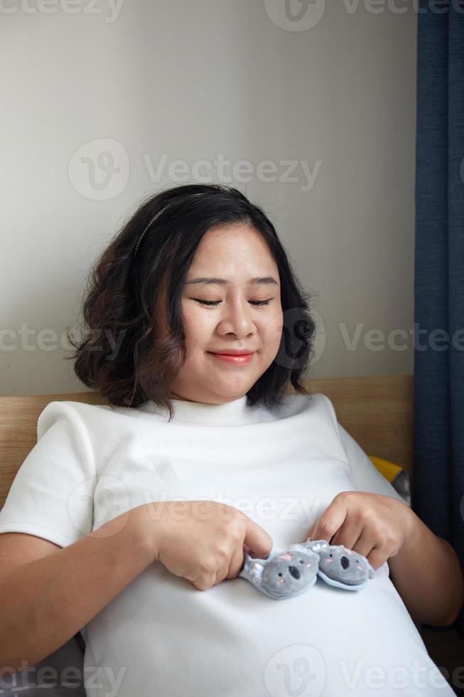 Young Happy Asian Pregnant holding small shoes for the unborn baby in her hand and look into the camera photo