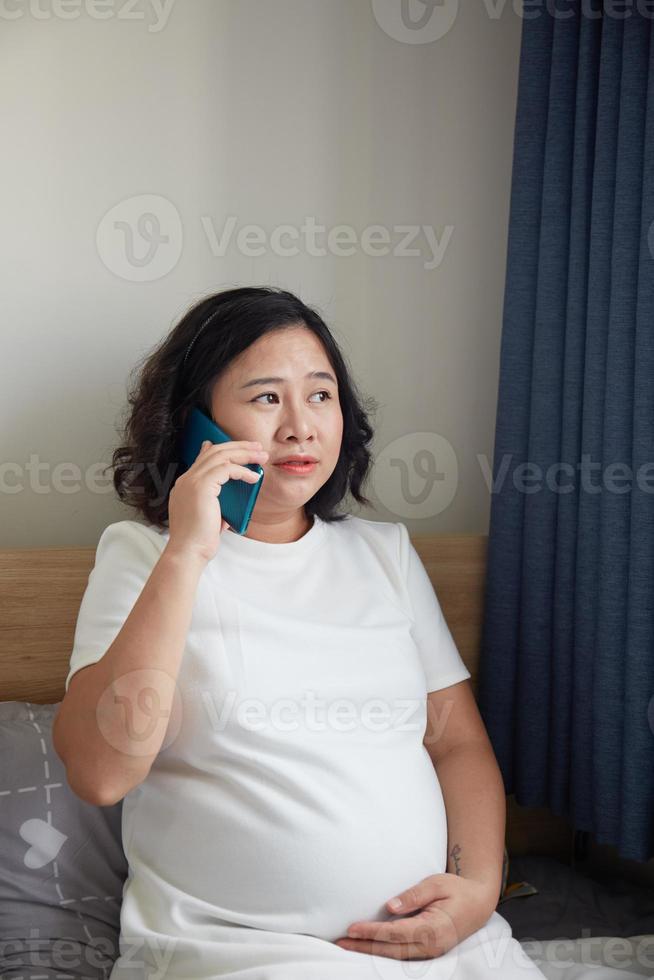 Young Asian Pregnant Woman standing near the window and using smart phone for checking information photo